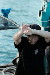 A person dressed in a black t-shirt is on a boat, shielding their face with one arm. In the background, there is water, suggesting a location near a body of water. A blue cloth is tied to the railing, and ropes are visible on the boat.