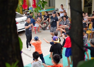 A group of children are enthusiastically participating in an outdoor activity, guided by adults. The scene includes a mix of children and adults, with the children engaged in movements and gestures as part of a group exercise or game. Adults are sitting around observing and some are interacting with the children. A car is parked nearby, and the setting appears to be a residential area or community space with chairs and miscellaneous items visible.