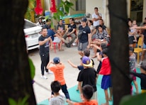 A group of children are enthusiastically participating in an outdoor activity, guided by adults. The scene includes a mix of children and adults, with the children engaged in movements and gestures as part of a group exercise or game. Adults are sitting around observing and some are interacting with the children. A car is parked nearby, and the setting appears to be a residential area or community space with chairs and miscellaneous items visible.