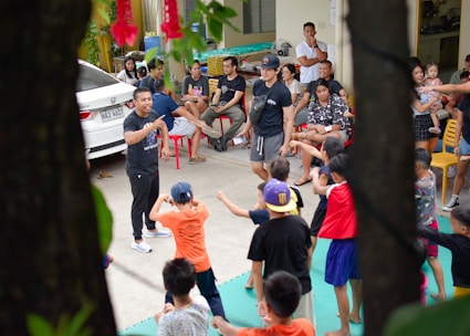 A group of children are enthusiastically participating in an outdoor activity, guided by adults. The scene includes a mix of children and adults, with the children engaged in movements and gestures as part of a group exercise or game. Adults are sitting around observing and some are interacting with the children. A car is parked nearby, and the setting appears to be a residential area or community space with chairs and miscellaneous items visible.