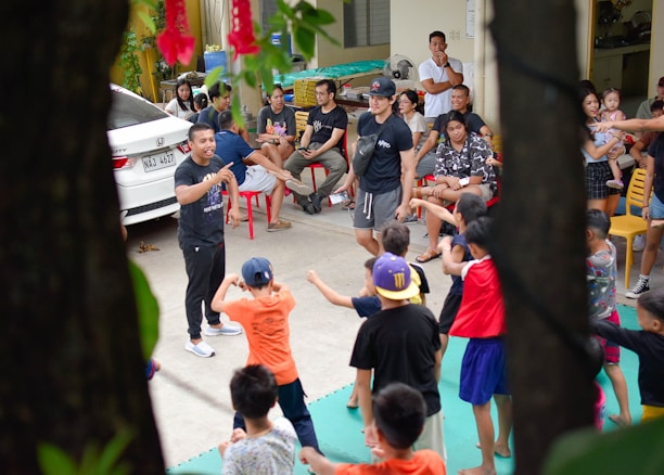 A group of children are enthusiastically participating in an outdoor activity, guided by adults. The scene includes a mix of children and adults, with the children engaged in movements and gestures as part of a group exercise or game. Adults are sitting around observing and some are interacting with the children. A car is parked nearby, and the setting appears to be a residential area or community space with chairs and miscellaneous items visible.