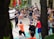 A group of children are enthusiastically participating in an outdoor activity, guided by adults. The scene includes a mix of children and adults, with the children engaged in movements and gestures as part of a group exercise or game. Adults are sitting around observing and some are interacting with the children. A car is parked nearby, and the setting appears to be a residential area or community space with chairs and miscellaneous items visible.