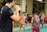 A group of children is practicing martial arts, focusing on their stance and positioning. An adult male instructor in a black shirt is facing them, likely giving instructions or demonstrating techniques. The setting appears to be indoors with various chairs and tables in the background.