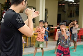 A group of children is practicing martial arts, focusing on their stance and positioning. An adult male instructor in a black shirt is facing them, likely giving instructions or demonstrating techniques. The setting appears to be indoors with various chairs and tables in the background.