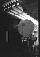 A black and white image showing an outdoor or studio photography setup, including a large umbrella-shaped softbox and a tripod stand. The background features structural elements like beams and a partially visible sign with text.