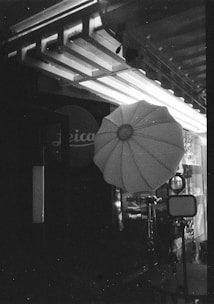 A black and white image showing an outdoor or studio photography setup, including a large umbrella-shaped softbox and a tripod stand. The background features structural elements like beams and a partially visible sign with text.