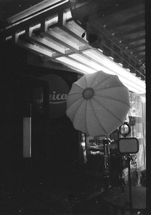 A black and white image showing an outdoor or studio photography setup, including a large umbrella-shaped softbox and a tripod stand. The background features structural elements like beams and a partially visible sign with text.
