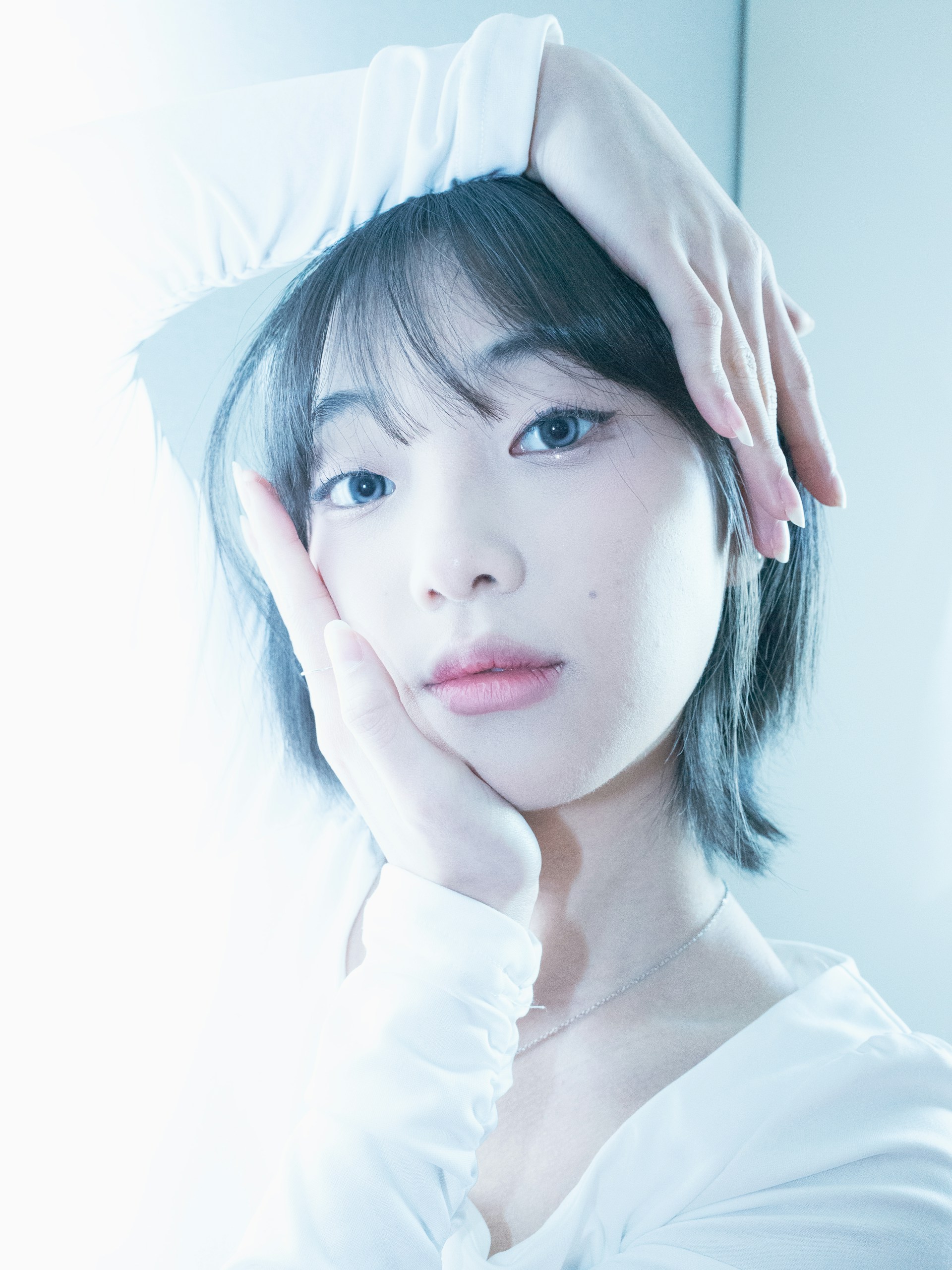 A person with short dark hair gazes directly at the camera, with both hands gently framing their face. The lighting is soft and ethereal, giving a serene atmosphere. The subject is wearing a white top, which enhances the calm and bright mood of the image.