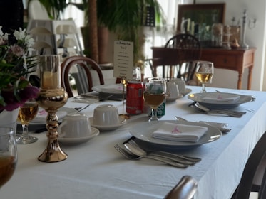 A dining table is elegantly set with white tablecloth and napkins. There are plates with cutlery neatly placed beside them, teacups with saucers, and glasses filled with a golden beverage. A menu or name card stands upright, and there are decorative elements like a gold candlestick and floral arrangements. In the background, wooden chairs and a sideboard with various objects are visible, alongside a potted plant.