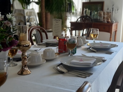 A dining table is elegantly set with white tablecloth and napkins. There are plates with cutlery neatly placed beside them, teacups with saucers, and glasses filled with a golden beverage. A menu or name card stands upright, and there are decorative elements like a gold candlestick and floral arrangements. In the background, wooden chairs and a sideboard with various objects are visible, alongside a potted plant.