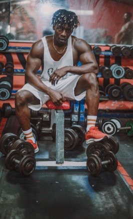 A muscular man is sitting on a bench in a gym, surrounded by dumbbells. He is wearing a white tank top, white shorts, and bright red shoes. The background includes a weight rack and mirrors, indicating a workout environment.