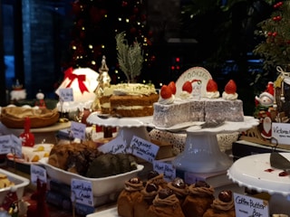 A selection of desserts and pastries displayed on a table, with a festive Christmas theme. Various cakes, cookies, and sweets are arranged on white plates and stands, some adorned with decorations such as strawberries. There are labels for each dessert including 'Coffee Butter Bar', 'Sweet Pumpkin Crumble', and 'Victoria'. The background features a decorated Christmas tree with lights and ornaments, adding a holiday ambiance.
