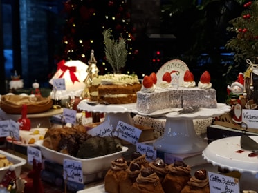 A selection of desserts and pastries displayed on a table, with a festive Christmas theme. Various cakes, cookies, and sweets are arranged on white plates and stands, some adorned with decorations such as strawberries. There are labels for each dessert including 'Coffee Butter Bar', 'Sweet Pumpkin Crumble', and 'Victoria'. The background features a decorated Christmas tree with lights and ornaments, adding a holiday ambiance.