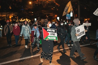 A group of people is participating in a nighttime protest or demonstration. They hold signs with messages related to supporting Palestine, and some individuals are wearing face masks. The environment is dimly lit, with streetlights and a few bright spots in the background, suggesting an urban setting.