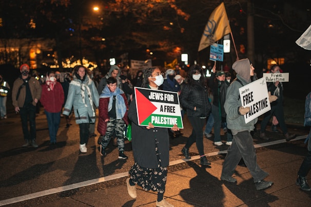 A group of people is participating in a nighttime protest or demonstration. They hold signs with messages related to supporting Palestine, and some individuals are wearing face masks. The environment is dimly lit, with streetlights and a few bright spots in the background, suggesting an urban setting.