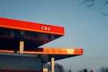 A modern gas station with a prominently displayed red sign reading 'CNG'. The structure features a clean, angular design with bright red and orange tones. The sky is clear with a gradient from light blue to pale orange near the horizon.