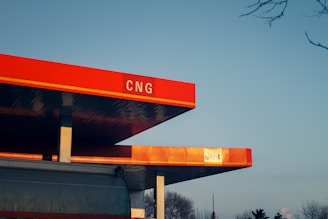 A modern gas station with a prominently displayed red sign reading 'CNG'. The structure features a clean, angular design with bright red and orange tones. The sky is clear with a gradient from light blue to pale orange near the horizon.
