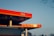 A modern gas station with a prominently displayed red sign reading 'CNG'. The structure features a clean, angular design with bright red and orange tones. The sky is clear with a gradient from light blue to pale orange near the horizon.