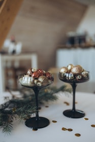 Two elegant black glass goblets are filled with assorted Christmas ornaments, primarily in gold, bronze, and silver colors. They rest on a white surface, surrounded by green pine branches and scattered golden confetti.