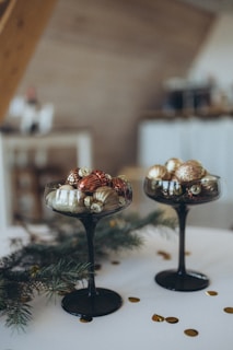 Two elegant black glass goblets are filled with assorted Christmas ornaments, primarily in gold, bronze, and silver colors. They rest on a white surface, surrounded by green pine branches and scattered golden confetti.