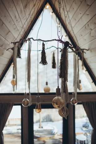 An interior view of an A-frame cabin displaying a rustic and artistic chandelier made from a wooden branch with hanging light bulbs and decorative tassels. The tall windows reveal a snowy landscape outside, adding to the cozy atmosphere.