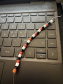 A close-up of a computer keyboard with Nordic/Scandinavian layout, including special characters such as Å, Æ, and Ø. On top of the keyboard lies a beaded wire adorned with small, colorful beads arranged in a pattern of black, white, red, and orange.