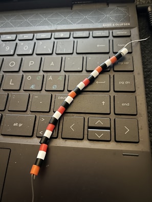 A close-up of a computer keyboard with Nordic/Scandinavian layout, including special characters such as Å, Æ, and Ø. On top of the keyboard lies a beaded wire adorned with small, colorful beads arranged in a pattern of black, white, red, and orange.