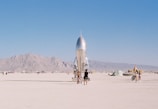 A shiny metallic rocket sculpture stands prominently in a desert landscape, surrounded by people walking and cycling. Distant mountains provide a backdrop under a clear blue sky.