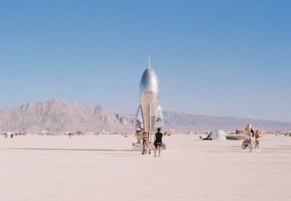 A shiny metallic rocket sculpture stands prominently in a desert landscape, surrounded by people walking and cycling. Distant mountains provide a backdrop under a clear blue sky.