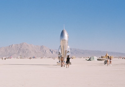 A shiny metallic rocket sculpture stands prominently in a desert landscape, surrounded by people walking and cycling. Distant mountains provide a backdrop under a clear blue sky.