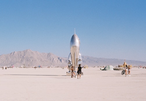 A shiny metallic rocket sculpture stands prominently in a desert landscape, surrounded by people walking and cycling. Distant mountains provide a backdrop under a clear blue sky.