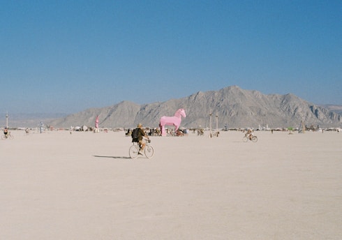 A vast, open desert landscape with scattered people riding bicycles. In the background, there is a large pink horse statue, surrounded by mountains under a clear blue sky.