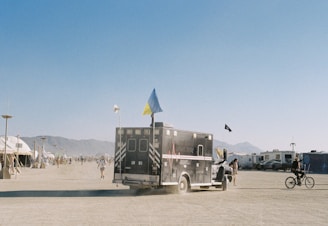 An outdoor scene in a desert-like area with an ambulance van adorned with flags driving on a dusty road. People are walking and riding bicycles around, and tents and RVs can be seen in the background, indicating a large gathering or festival setting. Distant mountains line the horizon under a clear blue sky.