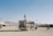An outdoor scene in a desert-like area with an ambulance van adorned with flags driving on a dusty road. People are walking and riding bicycles around, and tents and RVs can be seen in the background, indicating a large gathering or festival setting. Distant mountains line the horizon under a clear blue sky.
