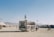An outdoor scene in a desert-like area with an ambulance van adorned with flags driving on a dusty road. People are walking and riding bicycles around, and tents and RVs can be seen in the background, indicating a large gathering or festival setting. Distant mountains line the horizon under a clear blue sky.