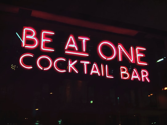A bright neon sign displays the words 'BE AT ONE COCKTAIL BAR' in red and pink colors against a dark background, suggesting a lively nightlife atmosphere.