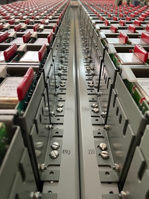 A close-up view down the center of a series of industrial electrical or communication panels, featuring rows of red tabs on each panel. Labeling and numbers are present on the metal surface, indicating organization and connectivity with screws and bolts as part of the assembly.
