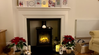 A cozy living room with a modern, electric fireplace centered on a white mantel. Decorative holiday cards and a small clock are displayed on the mantel. Poinsettia plants with vibrant red flowers flank the fireplace, adding a festive touch. There are candlesticks and a lit lantern placed around the hearth. A radiator is mounted on the wall, and a wicker basket with books is located near a beige sofa.
