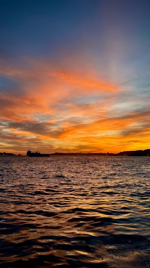 The image captures a sunset over the ocean, with vibrant orange and yellow hues in the sky reflecting off the water. Silhouettes of boats and land appear in the distance.