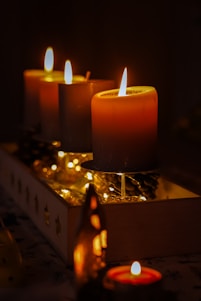 A warm, atmospheric setting featuring several lit candles arranged in a wooden box. The soft glow of the candle flames illuminates nearby pinecones and decorative string lights, creating an inviting and cozy ambiance.
