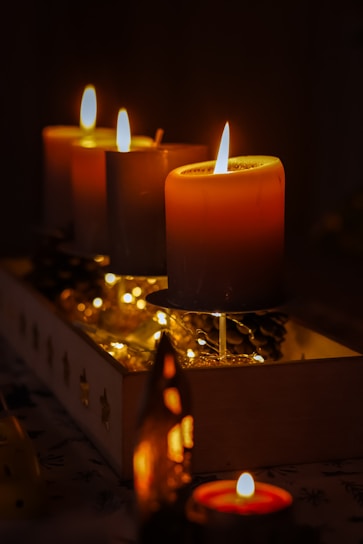 A warm, atmospheric setting featuring several lit candles arranged in a wooden box. The soft glow of the candle flames illuminates nearby pinecones and decorative string lights, creating an inviting and cozy ambiance.