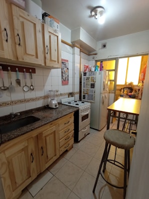 A narrow kitchen featuring wooden cabinets, a granite countertop, and various hanging utensils. The room includes a gas stove, a blender, and a refrigerator adorned with decorative magnets. There is a small window at the end of the space, allowing some natural light. A stool is positioned next to a tall table.