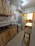 A narrow kitchen featuring wooden cabinets, a granite countertop, and various hanging utensils. The room includes a gas stove, a blender, and a refrigerator adorned with decorative magnets. There is a small window at the end of the space, allowing some natural light. A stool is positioned next to a tall table.