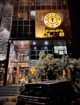 A modern building with a large illuminated sign for Gold's Gym Alexandria Club prominently displayed. The facade features glass windows and stone elements. In front, there is a neon sign of a muscular figure, some greenery, and a Range Rover is parked in the foreground.