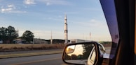 A tall, white and black rocket stands against a clear blue sky, surrounded by trees and buildings. An American flag is visible beside it. The image is taken from inside a vehicle, with the rocket visible through the side mirror as well, showing a reflection of the road and surrounding landscape.