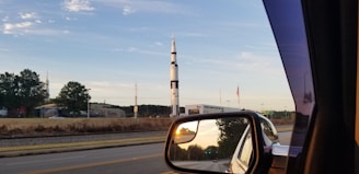 A tall, white and black rocket stands against a clear blue sky, surrounded by trees and buildings. An American flag is visible beside it. The image is taken from inside a vehicle, with the rocket visible through the side mirror as well, showing a reflection of the road and surrounding landscape.