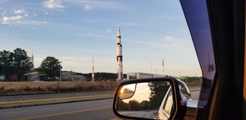 A tall, white and black rocket stands against a clear blue sky, surrounded by trees and buildings. An American flag is visible beside it. The image is taken from inside a vehicle, with the rocket visible through the side mirror as well, showing a reflection of the road and surrounding landscape.