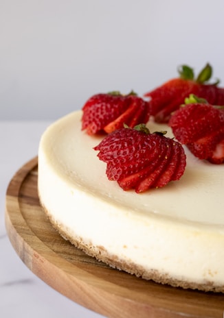 A creamy cheesecake is topped with sliced fresh strawberries and placed on a wooden board. The smooth texture of the cheesecake contrasts with the vibrant red strawberries. The edge of the board adds a rustic touch to the presentation.