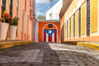 A narrow cobbled street flanked by colorful buildings with balconies and potted plants leads to a prominent building featuring a large Puerto Rican flag painted on its door.