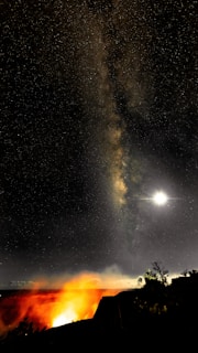 A starry night sky featuring the Milky Way galaxy stretches above a glowing volcanic landscape. The bright light of the moon illuminates part of the scene, casting a serene yet dramatic atmosphere.